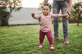 A toddler taking their first steps outside, with their arms outstretched for balance, with support from a parent or guardian.