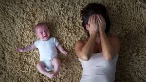 An overwhelmed mother lying on the floor, covering her face, with her crying baby beside her.