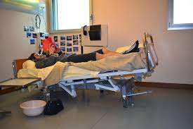 A person lying in bed in a Trendelenburg position, with their head lower than their feet, reading a book. The individual appears relaxed and focused on the book, with soft lighting illuminating the scene. The bed is set up with pillows for comfort, and the room has a calm, cozy atmosphere.
