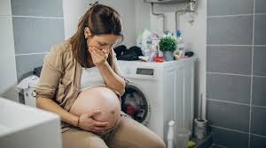 A woman in apparent labor sitting beside a birthing tub, visibly focused and breathing deeply. She is in a relaxed, seated position for comfort. The setting is a calm, soothing room with soft lighting, designed for a natural birthing experience.