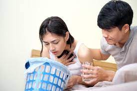 woman vomiting in a basket while her partner offers her water