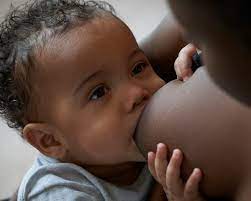 A baby nursing from their mother, making eye contact with a calm expression.