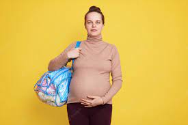A pregnant woman carrying a hospital bag, standing against a bright yellow background. She is wearing comfortable clothing, with a focused yet calm expression. The hospital bag is slung over her shoulder, and her baby bump is visible, emphasizing her pregnancy. The yellow background adds warmth and energy to the scene.