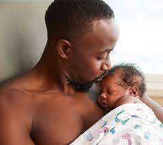 A father lovingly nestled with his baby in skin-to-skin contact, both wrapped in a soft blanket. The father holds the baby close to his chest, creating a tender, intimate moment. The scene is warm and peaceful, with soft lighting emphasizing the connection between them.
