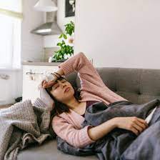 A sick woman lying on a couch, wrapped in a blanket. She has a tired expression, with a tissue box and a cup of tea nearby, suggesting she's unwell and resting in a cozy living room setting.