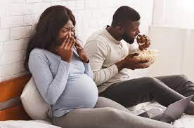 A pregnant woman sitting next to her partner, who is eating a burger. The woman looks uncomfortable, covering her nose with her hand as the smell of the food seems to make her feel nauseous. Her partner is focused on eating, unaware of her reaction. The setting is casual, with a neutral background.