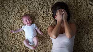 Overwhelmed woman laying on the floor next to a crying baby while covering her face