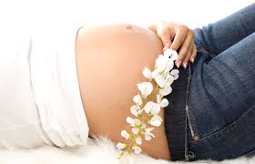 A first-trimester pregnant woman resting on her back, encircled by vibrant flowers, her exposed belly symbolizing new life.