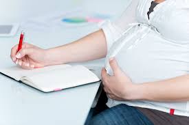 A pregnant woman sitting comfortably, journaling in a notebook. She has a serene expression as she writes, with her hand resting on her rounded belly. The setting is cozy, with soft lighting and a calm atmosphere, reflecting a peaceful moment of self-reflection during pregnancy.
