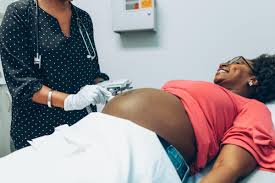 A healthcare professional standing beside a pregnant woman wearing a pink top, who is lying on an examination table with her abdomen uncovered. The healthcare professional is using a stethoscope to auscultate the fetal heart tones, while the pregnant woman looks relaxed and comfortable during the examination.