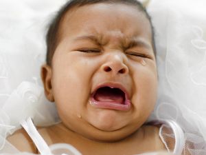 A baby with tears in their eyes, crying. The baby’s face shows distress, with scrunched features and a wide-open mouth. The image highlights the emotional expression of the baby, with soft lighting emphasizing the vulnerability of the moment. 