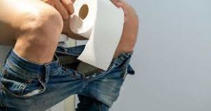 A person sitting on a toilet, holding a roll of toilet paper in their hand. The setting appears to be a clean and well-lit bathroom, with visible tiles and a neutral background.