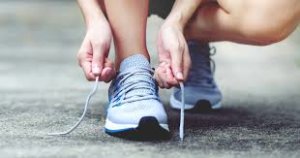A person bending down to tie their shoe laces. Their hands are focused on the task, with one lace held in each hand as they create a knot. The shoes are clearly visible, and the background may include neutral surroundings such as a floor or sidewalk. The image conveys an everyday, simple action of preparing to walk or run.