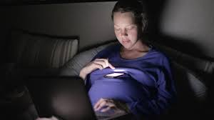 A woman lying in bed in a dimly lit room, illuminated by the soft glow of her phone screen. She is focused on the phone, her face gently lit in contrast to the dark surroundings.