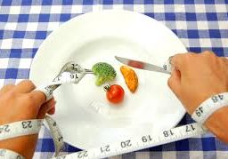 Person with hands tied with tape measure to plate with vegetables, holding spoon and fork: Fad diets and portion control.