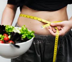 Woman measuring her abdomen with a measuring tape, holding a bowl of salad